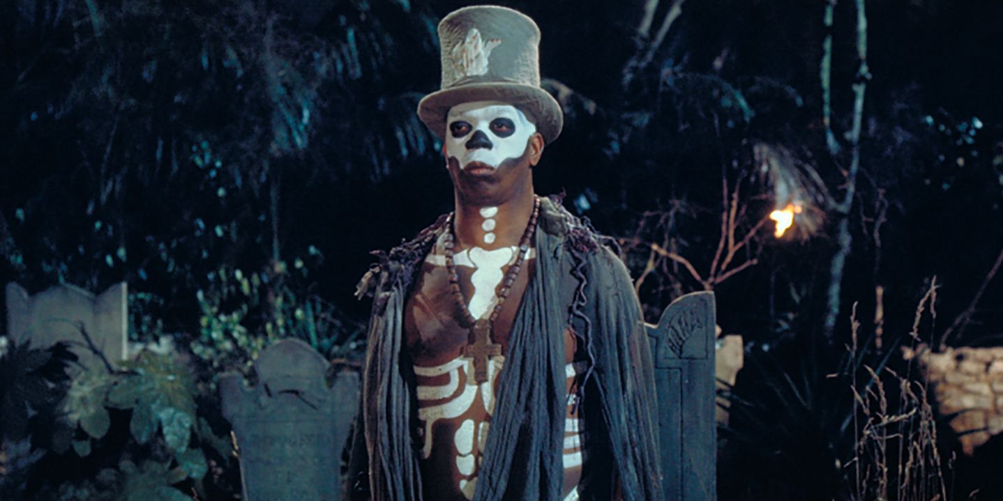 Baron Samedi orné de peinture squelette vaudou et d'un chapeau haut de forme se tient devant un cimetière dans 'Live and Let Die'