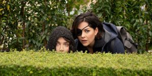 Eve Hewson and Sarah Greene peering over a bush