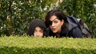 Eve Hewson and Sarah Greene peering over a bush