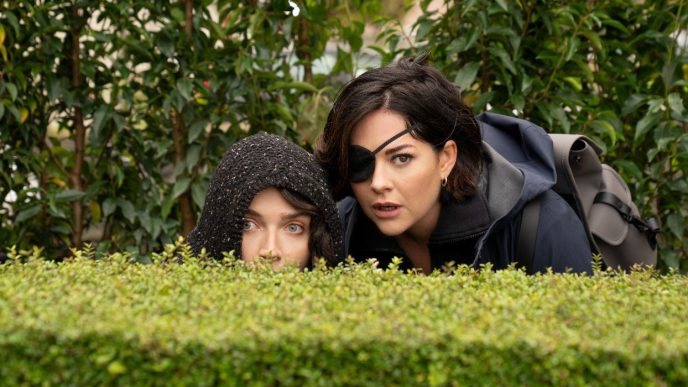 Eve Hewson and Sarah Greene peering over a bush