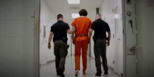 two police officers walk a prisoner dressed in orange down a corridor