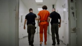 two police officers walk a prisoner dressed in orange down a corridor